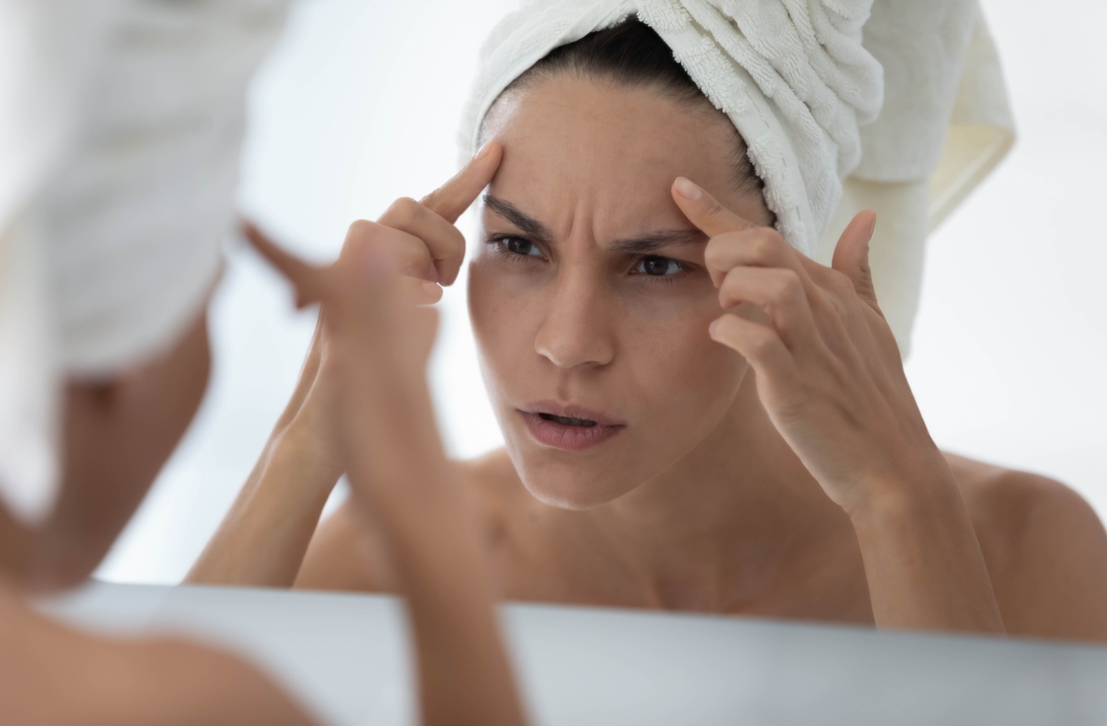 A frustrated woman with a towel on her head examining her forehead lines.