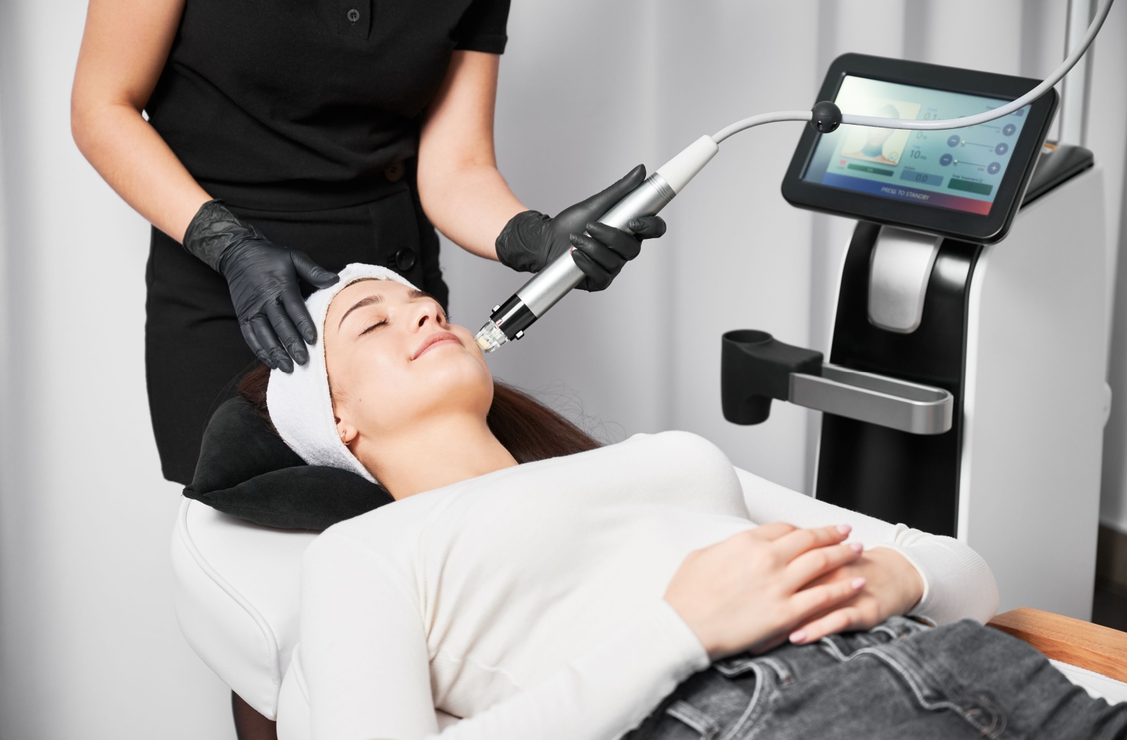 A person lies back with eyes closed while a clinician uses a handheld RF microneedling device.