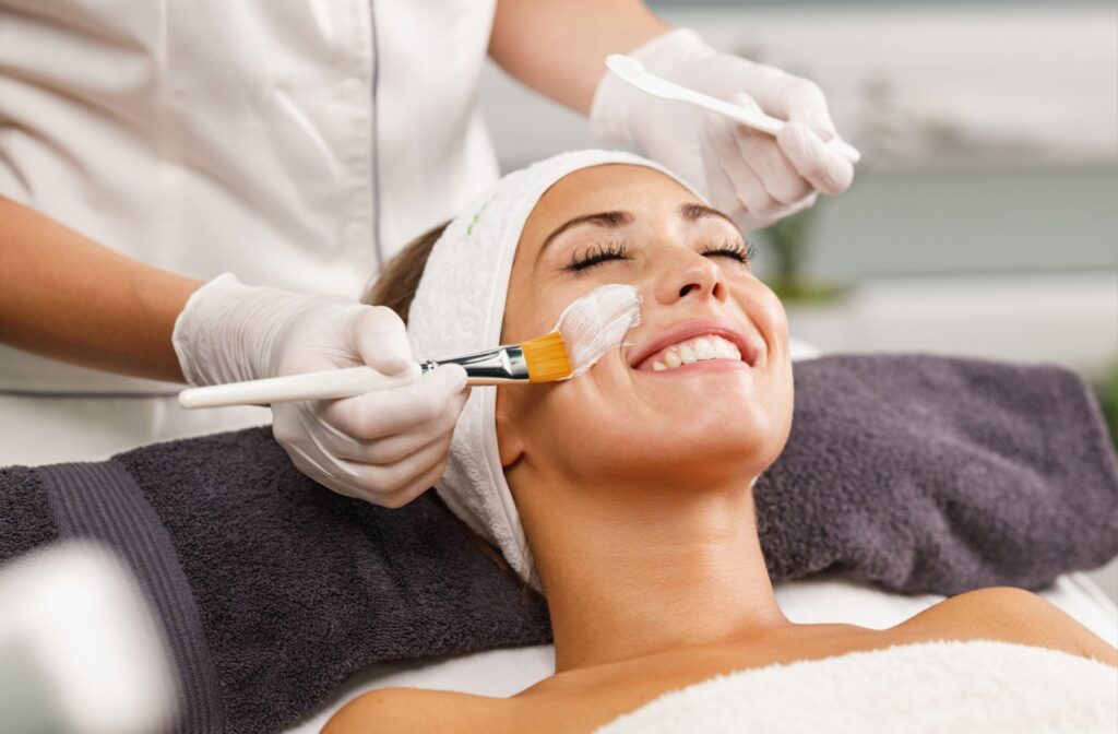 A person smiles while an esthetician applies a numbing cream with a brush.