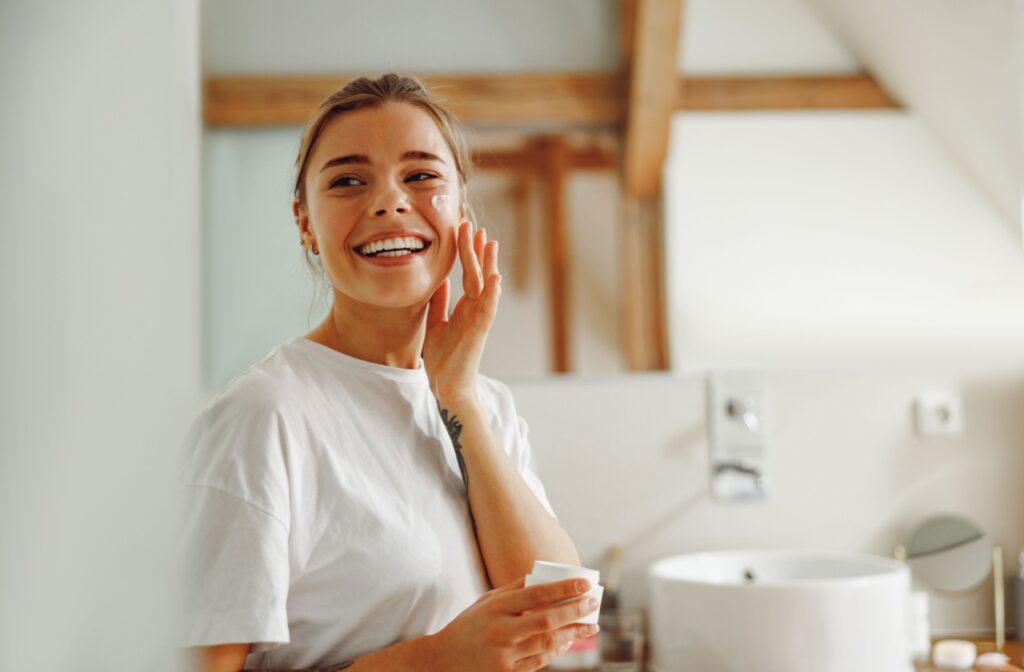 Woman smiling while applying moisturizer to her face in a bright bathroom, representing healthy skin and consistent skincare routine.