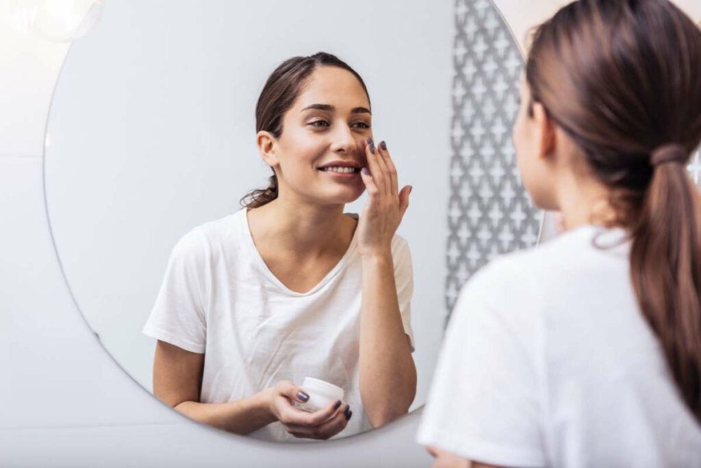 Smiling woman in white shirt applying facial cream while looking at reflection in round bathroom mirror holding moisturizer jar.