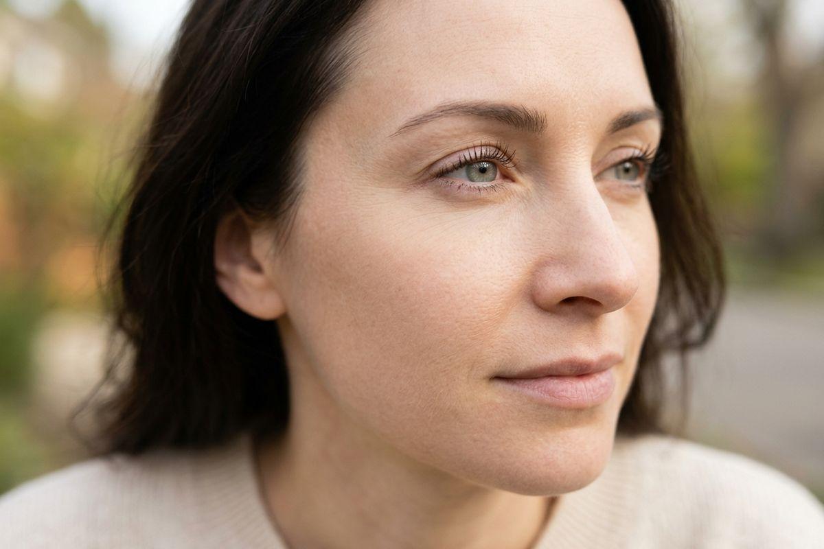 Close-up portrait of an adult with dark hair and green eyes gazing away, blurred outdoor background.